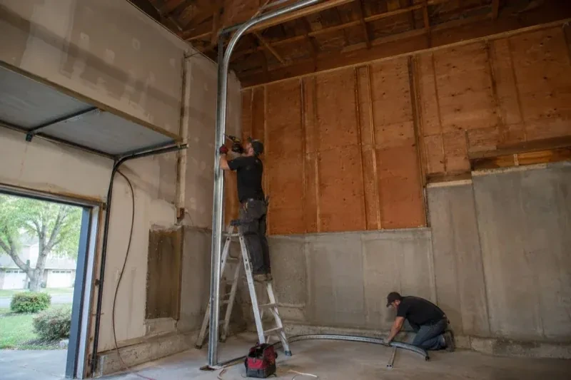 One person on a stepladder working beside a tall curved garage-door track while another kneels beside loose track sections in an unfinished garage. — High-Lift Garage Door Conversion, Thorold